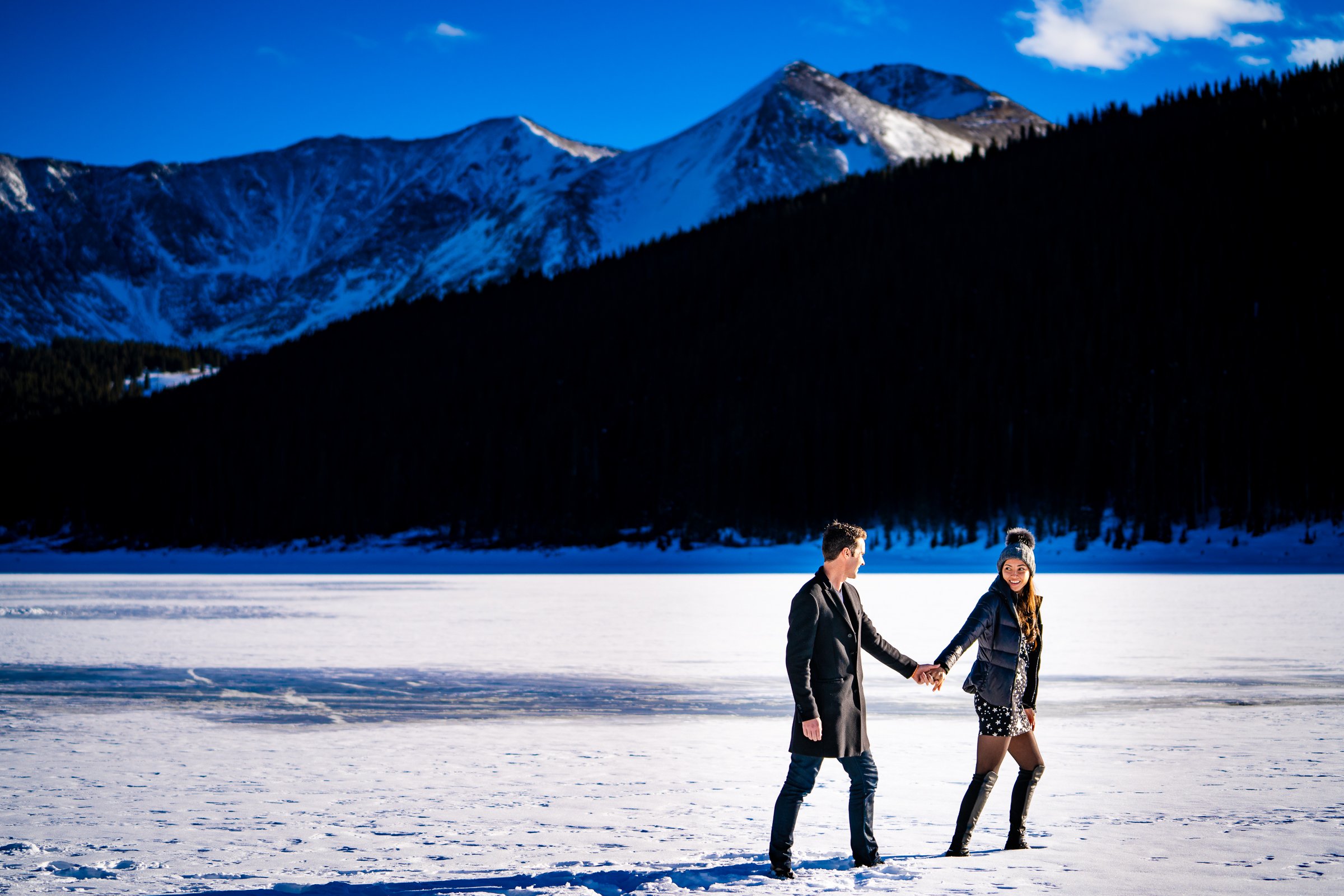 Engaged couple walks while holding hands on a frozen lake with snowcapped mountains in the background, Engagement Session, Engagement Photos, Engagement Photos Inspiration, Engagement Photography, Engagement Photographer, Winter Engagement Photos, Proposal Photos, Proposal Photographer, Proposal Photography, Winter Proposal, Mountain Proposal, Proposal Inspiration, Summit County engagement session, Summit County engagement photos, Summit County engagement photography, Summit County engagement photographer, Summit County engagement inspiration, Colorado engagement session, Colorado engagement photos, Colorado engagement photography, Colorado engagement photographer, Colorado engagement inspiration, Clinton Gulch Engagement